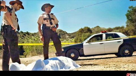 Two sheriffs in uniform stand near white-sheet-covered body at desert crime scene by patrol car