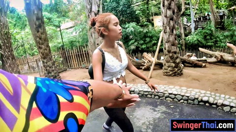 Thai girlfriend in tight pants and bralette walks in outdoor park holding bamboo stick pulled by hand amateur
