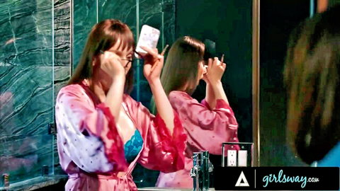 Two busty brunettes in pink satin robes and lingerie pose seductively taking mirror selfies in bathroom