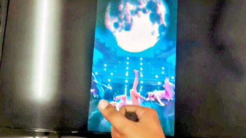 Hand holding phone screen showing naked women with tits and spread legs performing in blue-lit porn stage