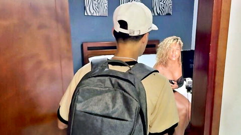 Asian guy with backpack approaches busty blonde exposing her huge bare tits on bedroom bed