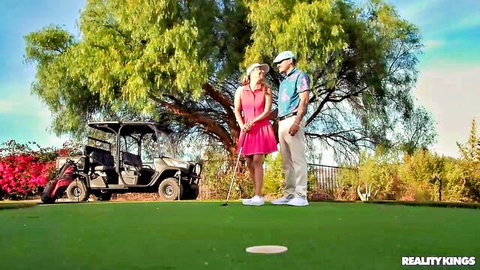 Summer Vixen in pink dress and hat poses with partner on sunny golf course