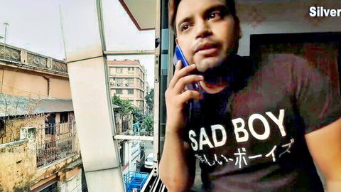 Muscular Indian man in Sad Boy t-shirt on phone call from balcony, teasing hotel room hookup