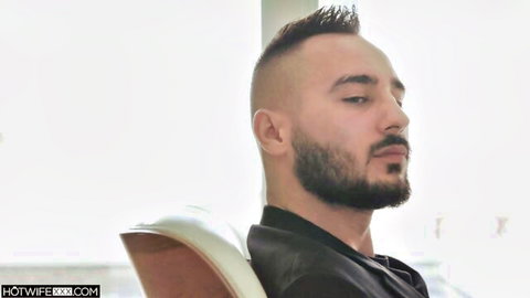 Handsome bearded man with mohawk haircut sits sideways in leather chair wearing black shirt