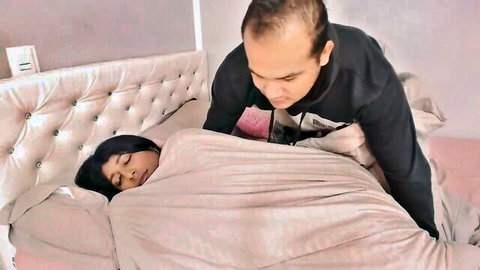 Balding man in black hoodie leans over sleeping Indian woman covered by gray blanket on pink bed