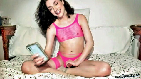 Smiling curly-haired TS in pink mesh lingerie sitting cross-legged on bed holding phone
