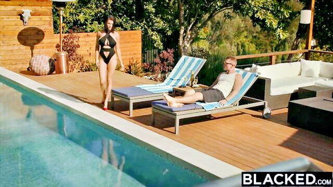 Busty tanned brunette in skimpy black bikini struts poolside toward lounging stud