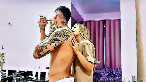 Tattooed muscular man smoking cigarette while blonde woman hugs his shirtless back in bedroom