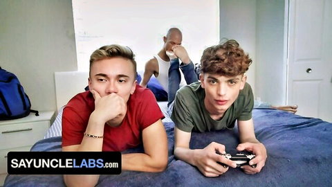 Young gay twinks in tank tops playing video games on bed with muscled Black daddy watching