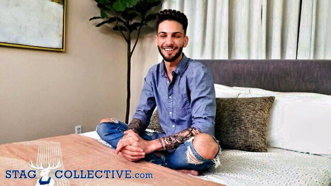 Muscular tattooed guy smiling cross-legged on bed in ripped jeans and shirt