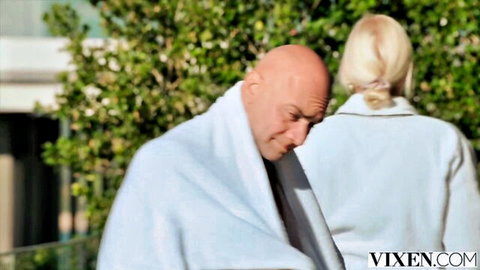 Bald man in white towel stares intently at blonde woman in robe from behind outdoors