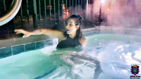 Curvy brunette in tight black swimsuit splashing excitedly in steamy outdoor hot tub at night