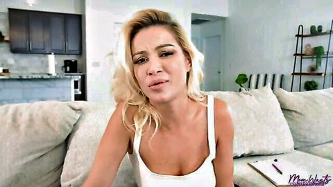 Blonde beauty in white tank top poses seductively on couch, cleavage tease