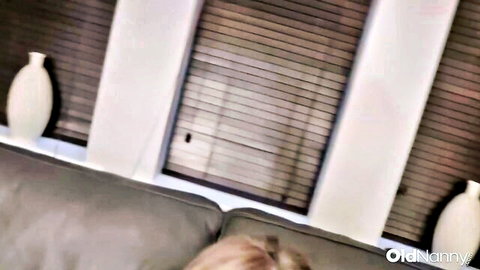 Blonde mature woman with long hair on gray couch by window blinds and vases