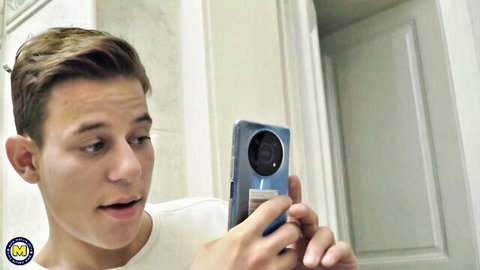 Young blonde guy in white t-shirt taking surprised selfie in bathroom mirror holding blue phone