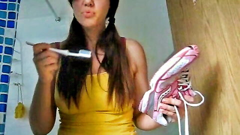 Skinny brunette in pigtails teases with toothbrush in mouth holding pink sneaker in yellow top
