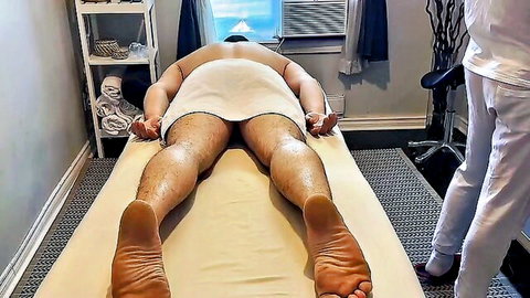 Hairy muscular man lying naked face down on massage table with tiny towel in steamy gay massage