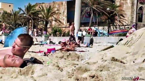 Smiling blond guy buried neck-deep in sand at crowded sunny beach with bikini-clad women lounging nearby