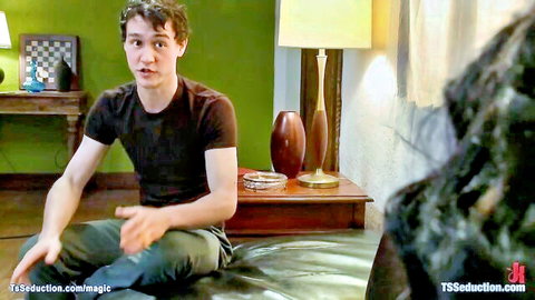 Young curly-haired brunette guy in black tee gesturing surprised in TS seduction bedroom