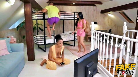 Busty ebony babe in skimpy orange top sits on floor gaming as Asian hottie in pink dress eyes guy hanging from bunk bed