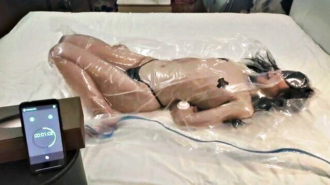 Blindfolded nude Asian woman tightly wrapped in clear plastic bondage exposing tits and pussy on bed