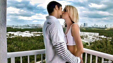 Skinny blonde babe in crop top passionately kissing muscular guy on sunny balcony overlooking city skyline