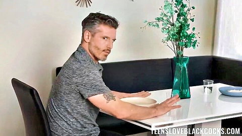 Mature gray-haired white man with beard and tattoo sitting at white table with green vase