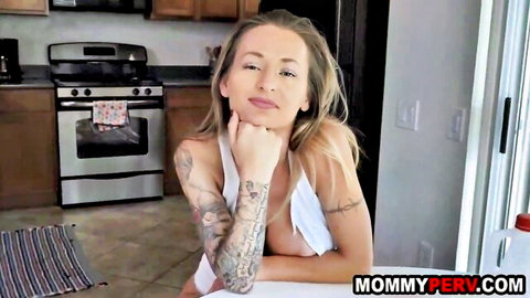 Tattooed blonde MILF with huge tits spilling from white tank top smiles in kitchen
