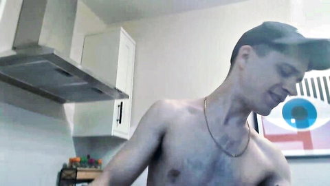Shirtless man smiling in kitchen, bare chest and necklace on cam