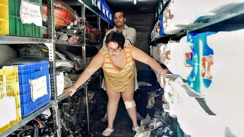 Chubby woman in striped top bending over in workshop as mechanic stands behind
