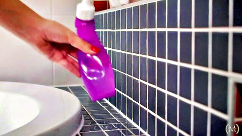Woman's hand holding purple spray bottle over bathroom sink with blue tiles