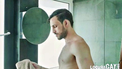 Muscular bearded gay hunk stands naked holding towel in steamy bathroom mirror