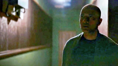 Bald man in dim green hallway, surprised expression, dark attire
