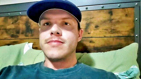Handsome mustached guy in backwards blue cap smiles teasingly in bed solo gay webcam