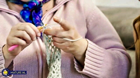 Mature granny crocheting yarn with hooked needle in pink hoodie and blue scarf
