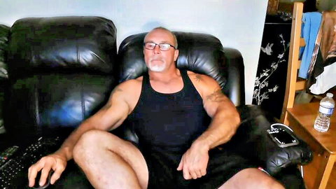 Muscular bald hairy man in tank top and shorts relaxing on couch for gay deepthroat webcam preview