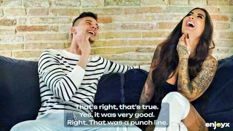 Tattooed brunette laughing with striped-shirt guy on couch in casual chat