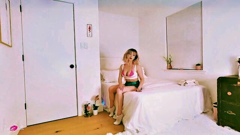 Hot blonde in pink bikini top and white shorts sits teasingly on bed with legs apart