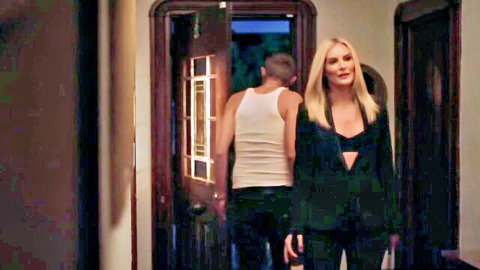 Seductive blonde in open black blazer and bra showing cleavage with guy in tank top by door