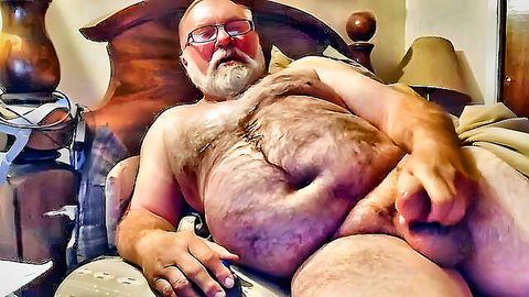 Hairy mature bear lying naked on bed stroking his thick cock