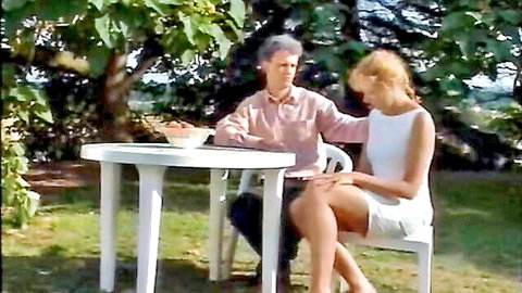 Blonde MILF in short white dress sits legs crossed with older man at outdoor table