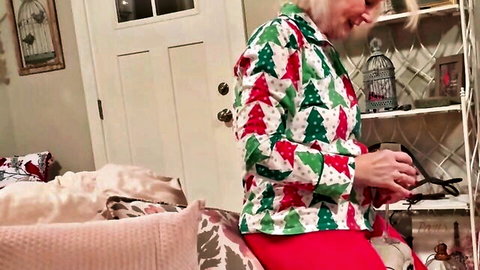 Mature GILF in sexy Christmas tree sweater and red pants holding scissors on bed teasing