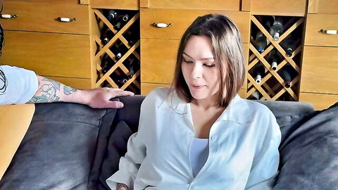 Tattooed man's hand gropes brunette's thigh as she sits on couch in open white shirt showing cleavage