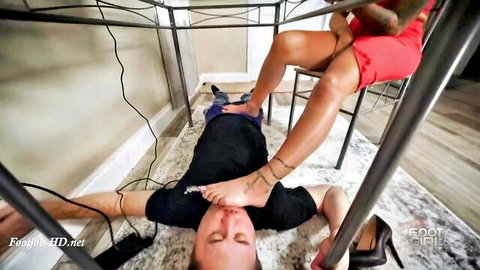 Tanned brunette in red dress presses bare foot on submissive man's face under kitchen table