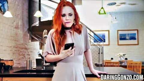Sexy redhead babe in low-cut gray dress showing cleavage while holding phone in kitchen