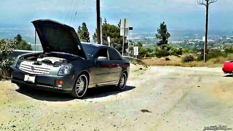 Black Cadillac with open hood broken down on dirt road near red car in sunny hills
