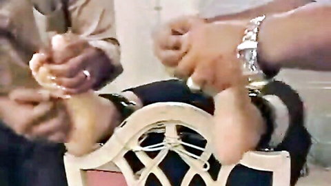 Two dark men fondle bare soles and toes of bound woman's spread feet in chair