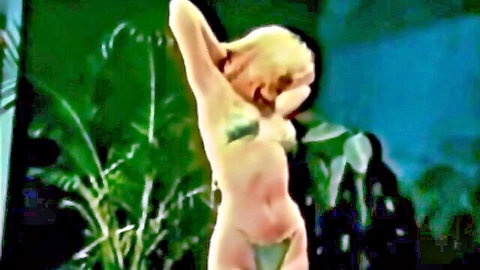 Vintage blonde in skimpy green bikini posing seductively with arms raised in tropical jungle
