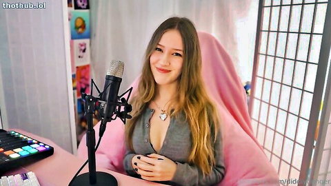 Cute blonde ASMR girl smiles in low-cut gray sweater showing deep cleavage at pink gaming mic