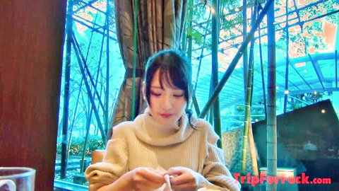 Cute Japanese teen in tight turtleneck sweater smiling at cafe table holding craft